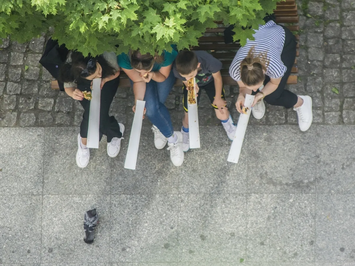Four people sharing long slices of pizza on a bench with a pigeon below